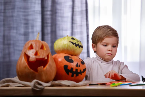 A preparar-se para o Halloween. O menino se senta em uma mesa com abóboras de Halloween na sala contra o fundo da janela. A criança prepara decorações para as férias. Fechar. — Fotografia de Stock