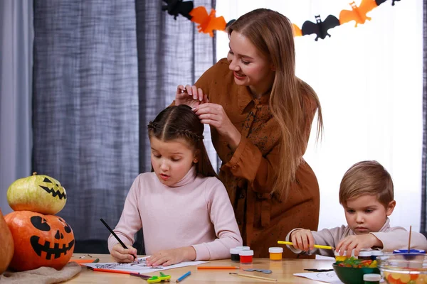 Família feliz em um quarto decorado para o Halloween. O menino brinca com tintas e lápis, a menina desenha, e neste momento sua mãe corrige o cabelo de suas filhas. A preparar-se para o Halloween. Fechar. — Fotografia de Stock