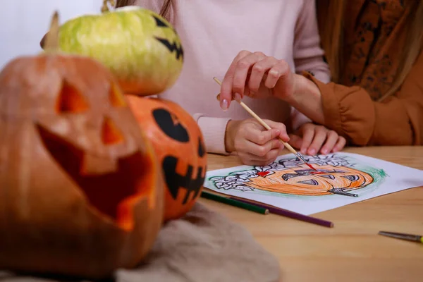 A família está se preparando para o Halloween e desenhando abóboras em papel com guache, sentadas à mesa. Close up de mãos de mãe e filha. — Fotografia de Stock