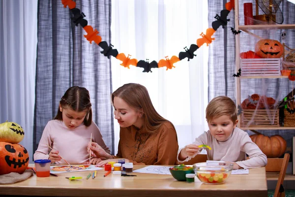 Mulher feliz com sua filha e filho se preparando para o Halloween. A família senta-se à mesa, a mãe ajuda a menina com o padrão de abóbora, e o menino procura doces. Fechar. — Fotografia de Stock