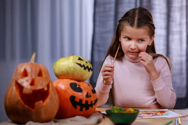 Uma menina adorável fez uma pausa de desenhar uma abóbora para comer doces guloseimas de Halloween. Uma criança em uma mesa em um quarto decorado para o Halloween. Fechar. — Fotografia de Stock
