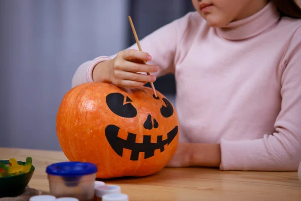 A menina prepara decorações para o Halloween em casa à mesa. Feche de uma mão de crianças que pinta uma cara assustadora em uma abóbora. Férias e conceito de decoração de Halloween. — Fotografia de Stock