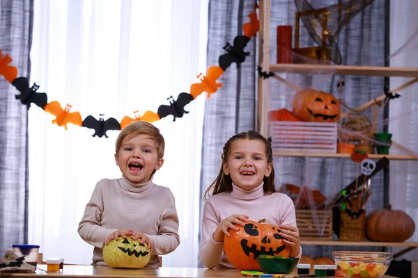 Irmão e irmã sentam-se à mesa com as mãos em suas abóboras de Halloween, que eles mesmos pintaram, sorrindo alegremente. As crianças adoram o Halloween. Fechar. — Fotografia de Stock