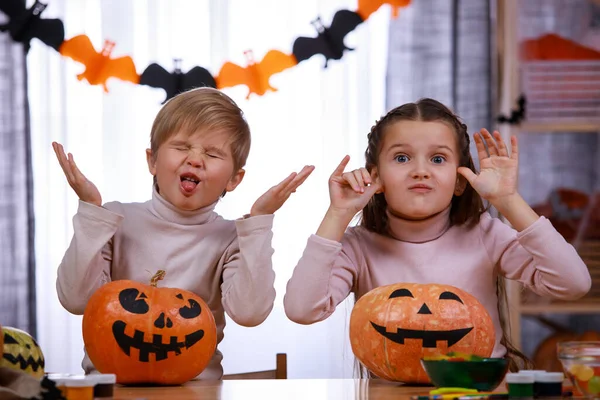 Duas crianças, um menino e uma menina, estão sentados em uma mesa perto de abóboras com rostos assustadores pintados e fazendo rostos. Irmão e irmã na sala de estar decorados para o Halloween. Fechar. — Fotografia de Stock