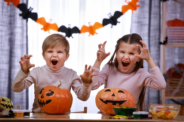 Duas crianças, um menino e uma menina, estão sentadas em uma mesa perto de abóboras de Halloween e fazem caras assustadoras pedindo guloseimas. Irmão e irmã na sala de estar decorados para o Halloween. Fechar. — Fotografia de Stock