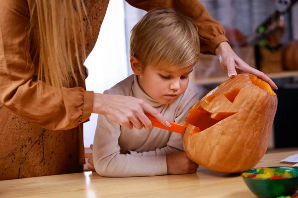 A mãe mostra ao filho como fazer uma cara de Lanterna Jack de Halloween na grande abóbora laranja. Feche de uma mão de mulher que esculpe uma cara terrível em uma abóbora. Conceito de decoração feriados. — Fotografia de Stock