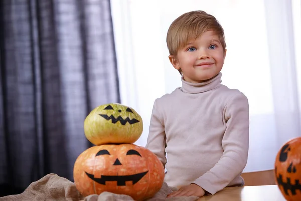 Um retrato de um menino adorável sentado em uma mesa com três abóboras com rostos assustadores pintados para o Halloween. As crianças adoram o Halloween. Fechar. — Fotografia de Stock