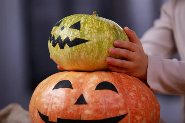 A preparar-se para o Halloween. Na mesa está uma abóbora com uma cara horrível pintada, o menino coloca outra em cima dela. Feche de uma mão de crianças com uma abóbora. Halloween.. — Fotografia de Stock