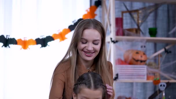 Jovem mulher bonita com longas tranças de cabelo as tranças de uma menina em um quarto decorado para o dia das bruxas, o fundo é turvo. A mãe toma conta da filha. Movimento lento. Fechar. — Vídeo de Stock