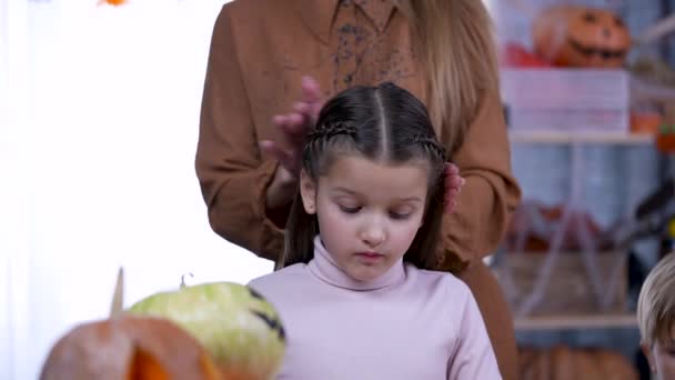 Dívka vedle chlapce se připravuje na Halloween ve zdobené místnosti s rozmazaným pozadím. Mladá žena v tuto chvíli opravuje dívčí vlasy. Zpomal. Zavřít. — Stock video