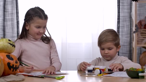 La niña está pintando un dibujo de una calabaza en la mesa de la habitación, y su hermano menor está jugando con pinturas cerca de ella. Halloween, diversión otoñal para niños. En cámara lenta. De cerca.. — Vídeos de Stock