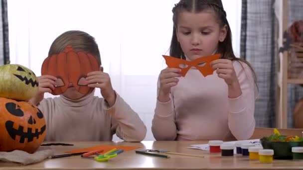 カボチャとバットの形をした紙から切り出されたフェイスマスクを試着している部屋のテーブルに兄と妹が座っています。子供たちはハロウィーンの準備を楽しんでいます。スローモーション。閉じろ!. — ストック動画