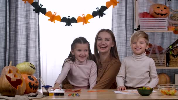 La familia se está divirtiendo preparándose para Halloween. Mamá, hija e hijo en una habitación decorada para las vacaciones están sentados a la mesa riendo y abrazándose. En cámara lenta. De cerca.. — Vídeo de stock