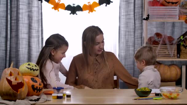 Lycklig mor kramar sina barn med kärlek. En kvinna lär sin son en speciell hälsning när hon sitter vid ett bord i ett rum inrett för Halloween. Sakta i backarna. Närbild. — Stockvideo