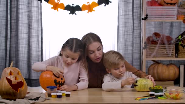 Mamma och hennes barn förbereder sig för Halloween. En pojke och en flicka ritar läskiga ansikten på pumpor, och deras mamma hjälper dem. Semester och Halloween dekoration koncept. Sakta i backarna. Närbild. — Stockvideo