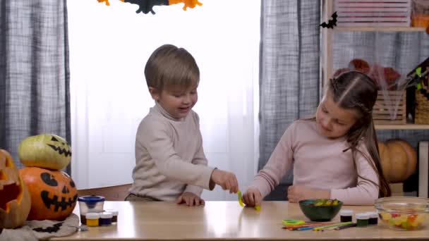 Un niño y una niña se sientan en una mesa con golosinas y calabazas en una habitación decorada para Halloween. Los niños juegan con gusanos gelatinosos. Halloween. En cámara lenta. De cerca.. — Vídeo de stock