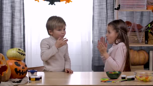 Een jongen en een meisje zitten aan een tafel met lekkernijen en pompoenen in een kamer versierd met Halloween. De broer plaagt zijn zus met een jelly worm en eet het zelf op. Langzame beweging. Sluiten.. — Stockvideo
