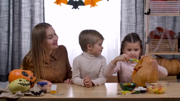 Family in a room decorated for Halloween are sitting at the table. A girl carves Jacks flashlight from a pumpkin, and her mother and her brother are watching her. Slow motion. Close up. — Stock Video