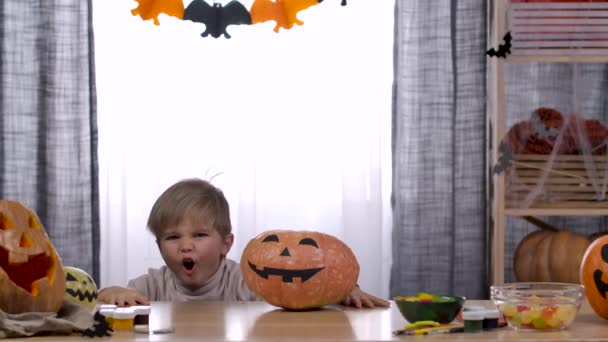 Em um quarto decorado para o Halloween, o menino se escondeu sob uma mesa com uma abóbora com um rosto assustador pintado nela. A criança salta de baixo da mesa e começa a assustar. Movimento lento. Fechar. — Vídeo de Stock