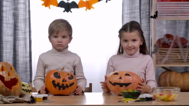 Két gyerek, egy fiú és egy lány, sütőtököket nevelnek előttük ijesztő arccal. Tököket tesznek az asztalra és arcokat vágnak. A gyerekek imádják a Halloweent. Lassú mozgás. Közelről.. — Stock videók