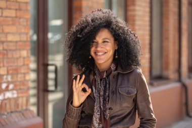 Afro-Amerikan genç bir kadının portresi tabelayı düzeltiyor ve gülümsüyor. Kahverengi deri ceketli esmer, bulanık tuğla binanın arka planında poz veren. Kapat..