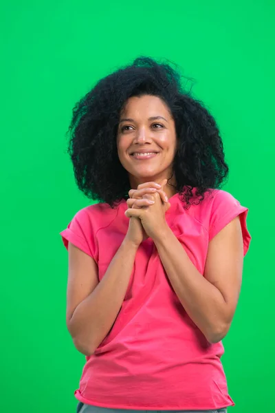 Portrett av unge, glade kvinnelige afroamerikanere som drømmer om noe hyggelig. En svart kvinne med krøllete hår i rosa skjorte poserer på en grønn skjerm i studioet. Lukk. – stockfoto