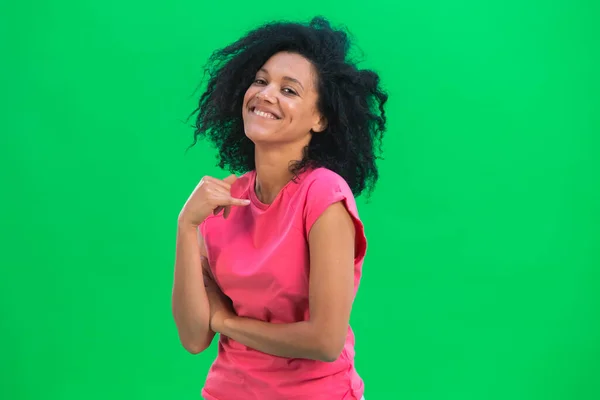 Portrett av unge kvinnelige afroamerikanske flørtete smil. En svart kvinne med krøllete hår i rosa skjorte poserer på en grønn skjerm i studioet. Lukk. – stockfoto