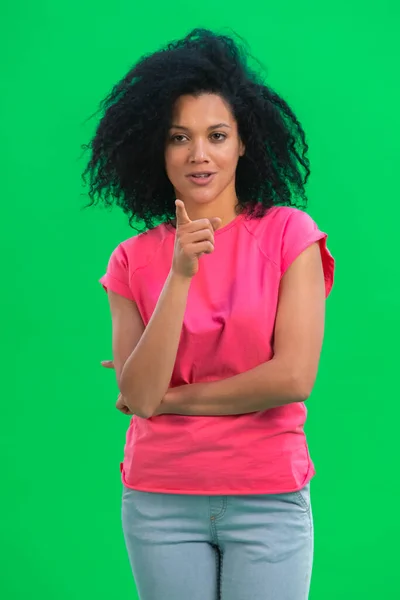 Muotokuva, jossa nuori afroamerikkalainen nainen osoittaa kameraa etusormella. Musta nainen kihara tukka vaaleanpunainen t paita aiheuttaa vihreällä ruudulla studiossa. Sulje se.. — kuvapankkivalokuva