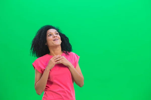 Portrett av unge kvinnelige afroamerikanere som ser seg rundt med utrolig glede og ømhet. En svart kvinne med krøllete hår i rosa skjorte poserer på en grønn skjerm i studioet. Lukk. – stockfoto