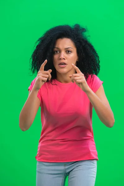 Portrait of young female African American looking at the camera with excitement. Black woman with curly hair in pink tshirt poses on green screen in the studio. Close up. — Stock Photo, Image