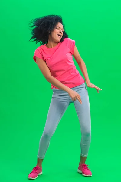 Portrait of young female African American cheerfully dancing the dance of victory, joy or good mood. Black woman with curly hair poses on green screen in studio. Full length. — Stock Photo, Image
