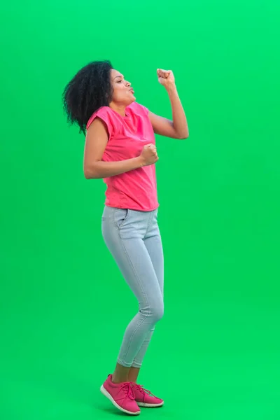 Portrait of young female African American cheerfully dancing the dance of victory, joy or good mood. Black woman with curly hair poses on green screen in studio. Full length. — Stock Photo, Image