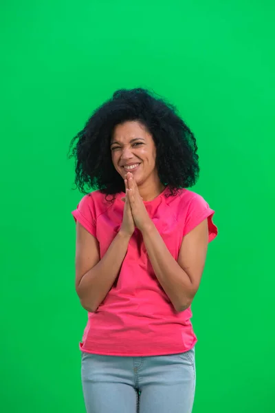 Nuoren afroamerikkalaisen naisen muotokuva, joka pitää kämmenet yhdessä ja pyytää jotain. Musta nainen kihara tukka vaaleanpunainen t paita aiheuttaa vihreällä ruudulla studiossa. Sulje se.. — kuvapankkivalokuva