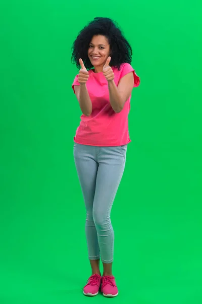 Retrato de jovem afro-americana mostrando os polegares para cima e sorrindo. Mulher preta com cabelo encaracolado em camiseta rosa e jeans posa na tela verde no estúdio. Comprimento total. — Fotografia de Stock