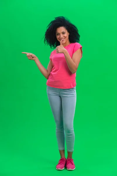 一个年轻的非洲裔美国女性的画像,用手尖着东西,并展示了她的大拇指.身穿粉色T恤和牛仔裤的卷发黑人妇女在工作室的绿色屏幕上摆出姿势。全长. — 图库照片