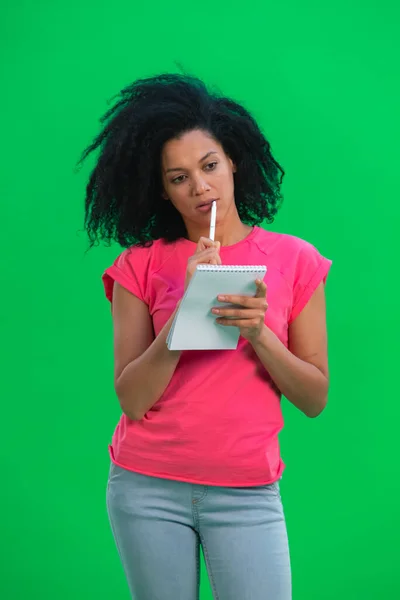 Portrett av ung kvinnelig afroamerikansk tenkning, deretter lykkelig skriving med penn i notatbok. En svart kvinne med krøllete hår poserer på en grønn skjerm i studioet. Lukk. – stockfoto