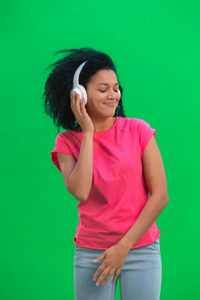 若い女性のアフリカ系アメリカ人のダンスの肖像と大きな白いヘッドフォンで音楽を楽しんでいます。ピンクのTシャツに巻き毛の黒い女がスタジオの緑のスクリーンにポーズをとっている。閉じろ!. — ストック写真