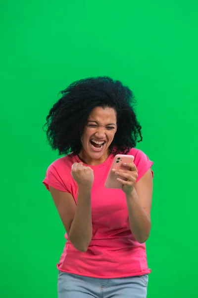 Retrato de una joven afroamericana está enviando mensajes de texto por teléfono y se regocijan con las buenas noticias. Mujer negra con el pelo rizado en camiseta rosa posa en la pantalla verde en el estudio. De cerca.. —  Fotos de Stock