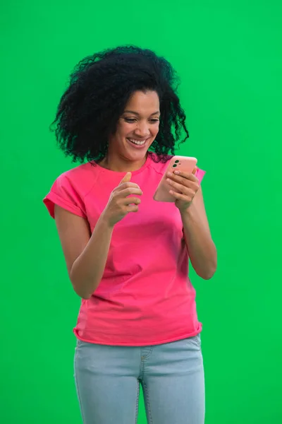 Retrato de una joven afroamericana está enviando mensajes de texto por teléfono y se regocijan con las buenas noticias. Mujer negra con el pelo rizado en camiseta rosa posa en la pantalla verde en el estudio. De cerca.. —  Fotos de Stock