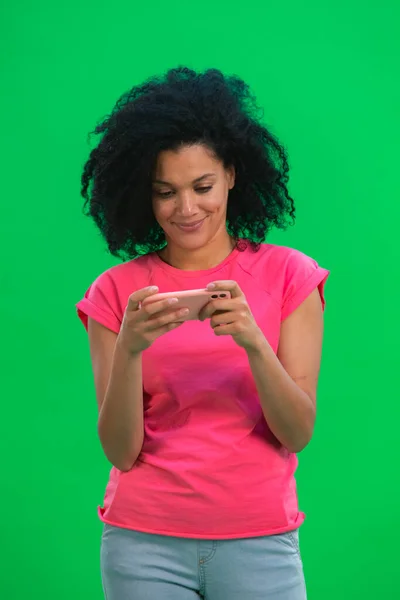 Retrato de una joven afroamericana jugando en un smartphone o enviando mensajes de texto. Mujer negra con el pelo rizado en camiseta rosa posa en la pantalla verde en el estudio. De cerca.. —  Fotos de Stock