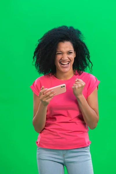 Portret młodej Afroamerykanki grającej na smartfonie i cieszącej się zwycięstwem. Czarna kobieta z kręconymi włosami w różowej koszulce pozuje w studio na zielonym ekranie. Zamknij się.. — Zdjęcie stockowe