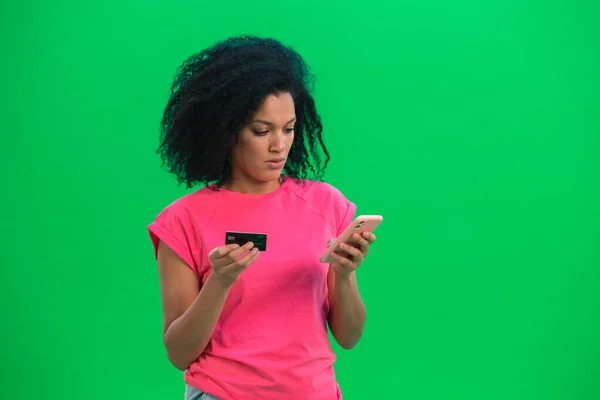 Retrato de una joven afroamericana que realiza una compra en línea con tarjeta de crédito y teléfono. Mujer negra con el pelo rizado en camiseta rosa posa en la pantalla verde en el estudio. De cerca.. —  Fotos de Stock