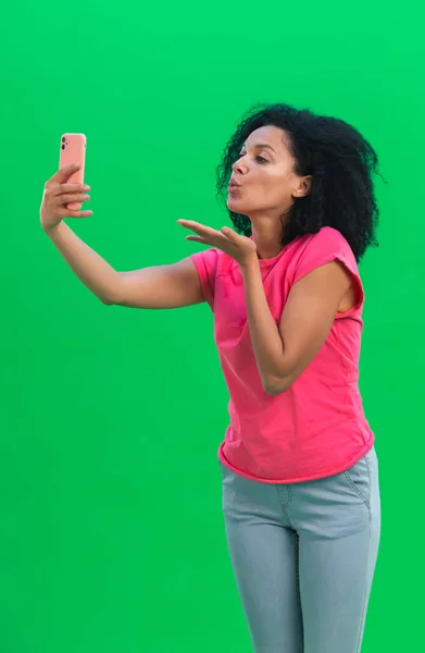 Portret młodej Afroamerykanki rozmawiającej na video czacie przez telefon komórkowy i dmuchającej pocałunek. Czarna kobieta z kręconymi włosami w różowej koszulce i dżinsach pozuje w studio na zielonym ekranie. Zamknij się. — Zdjęcie stockowe