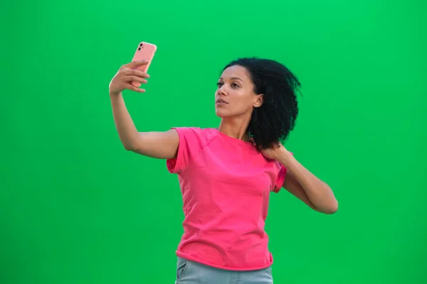 携帯電話で自撮りをする若い女性アフリカ系アメリカ人の肖像画。ピンクのTシャツに巻き毛の黒い女がスタジオの緑のスクリーンにポーズをとっている。閉じろ!. — ストック写真