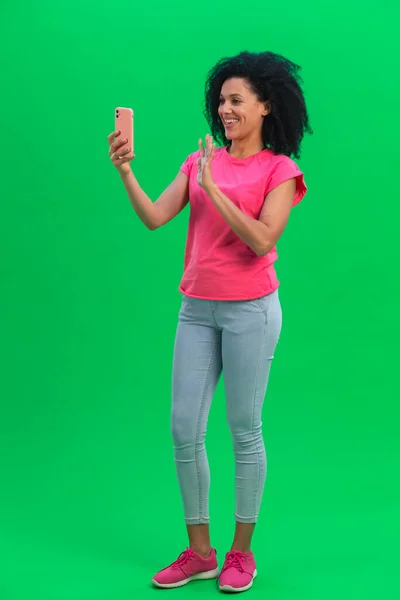 Портрет молодої афроамериканки, яка розмовляє на відео, користується мобільним телефоном і радіє. Чорна жінка з кучерявим волоссям в рожевій сорочці і джинсах пози на зеленому екрані в студії. Повна довжина. — стокове фото