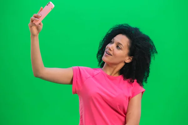 Nuoren afroamerikkalaisen naisen muotokuva, joka tekee selfien matkapuhelimella. Musta nainen kihara tukka vaaleanpunainen t paita aiheuttaa vihreällä ruudulla studiossa. Sulje se.. — kuvapankkivalokuva