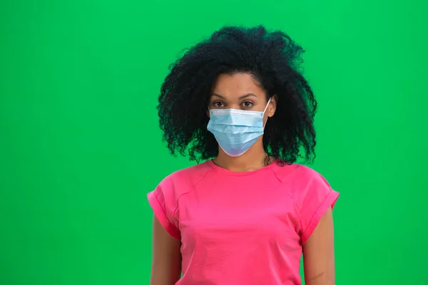 Portret młodej Afroamerykanki patrzącej w kamerę w masce medycznej. Czarna kobieta z kręconymi włosami w różowej koszulce pozuje w studio na zielonym ekranie. Zamknij się.. — Zdjęcie stockowe