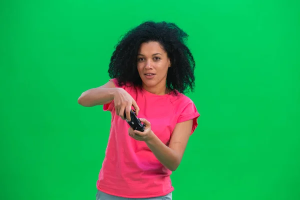 Muotokuva nuoresta afroamerikkalaisesta naisesta pelaamassa videopeliä langattoman ohjaimen avulla. Musta nainen kihara tukka vaaleanpunainen t paita aiheuttaa vihreällä ruudulla studiossa. Sulje se.. — kuvapankkivalokuva