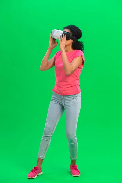 Portrait of young female African American with virtual reality headset or 3d glasses . Black woman with curly hair in pink tshirt and jeans poses on green screen in the studio. Full length. — Stock Photo, Image