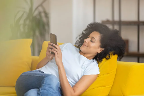 Retrato de una joven afroamericana enviando mensajes de texto o viendo información en su teléfono. Morena con el pelo rizado sentado en un sofá amarillo en una habitación luminosa en casa. De cerca.. — Foto de Stock